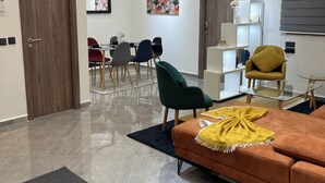 Una sala de estar moderna con un sofá marrón, una manta amarilla y un sillón verde. Se observa un comedor con una mesa y sillas al fondo.