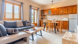 Uma sala de estar moderna com sofá e mesa de centro, cozinha com armários de madeira e geladeira, e grandes janelas com cortinas.