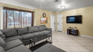 A living room with a grey sectional sofa, a small black coffee table, a flat-screen TV on a wooden stand, and a door in the background.