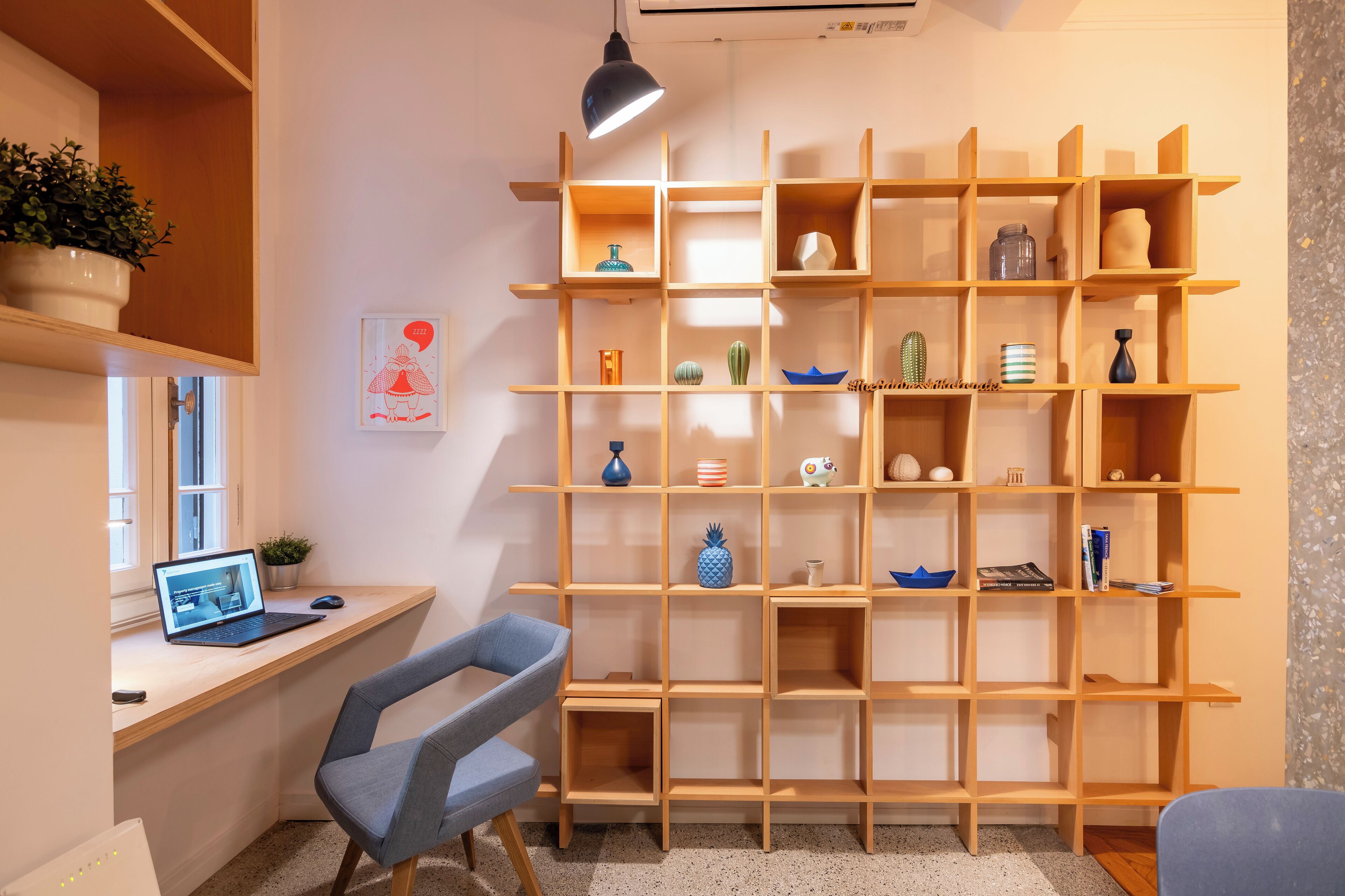 Un espace de travail moderne avec un bureau en bois, un ordinateur portable et une chaise bleue. Une étagère en bois, ornée de divers objets décoratifs, se trouve à l’arrière-plan.