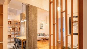 Moderni olohuone, jossa on puinen ruokapöytä, tuoleja, sohva ja sohvapöytä. Huoneessa on myös kirjahylly, jossa on erilaisia esineitä, sekä riippuvalaisin.