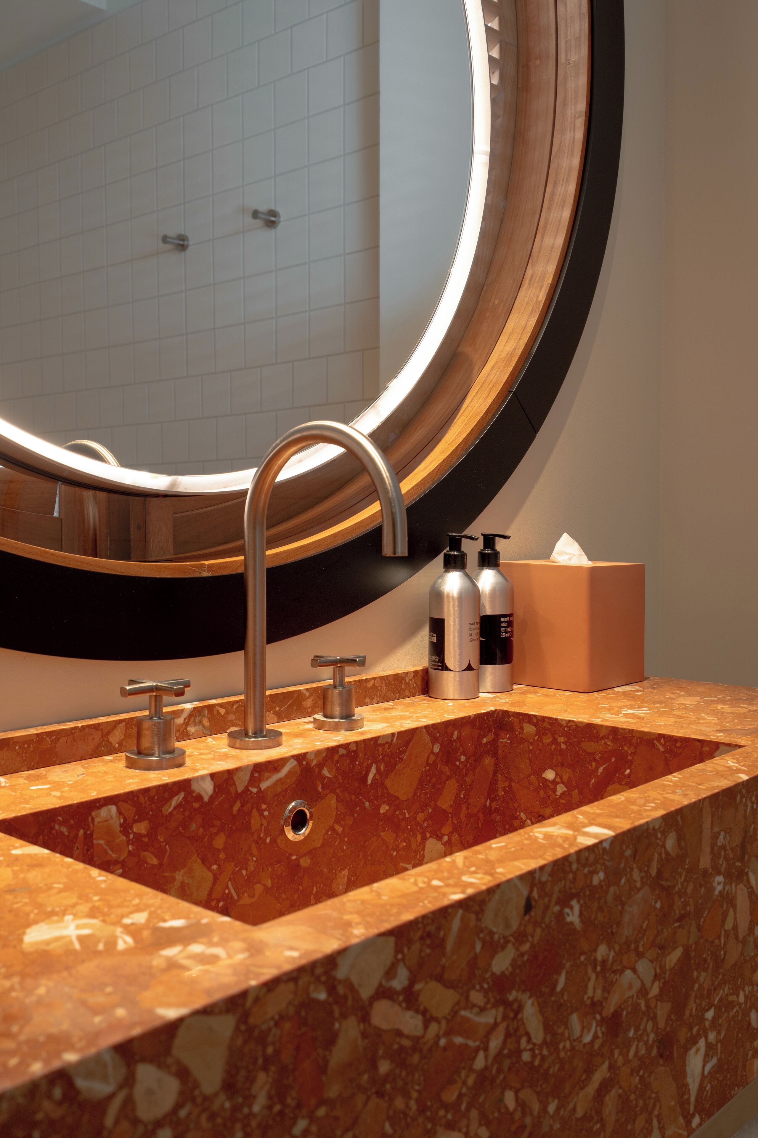 A bathroom with a marble sink, a round mirror, and two bottles of shampoo.