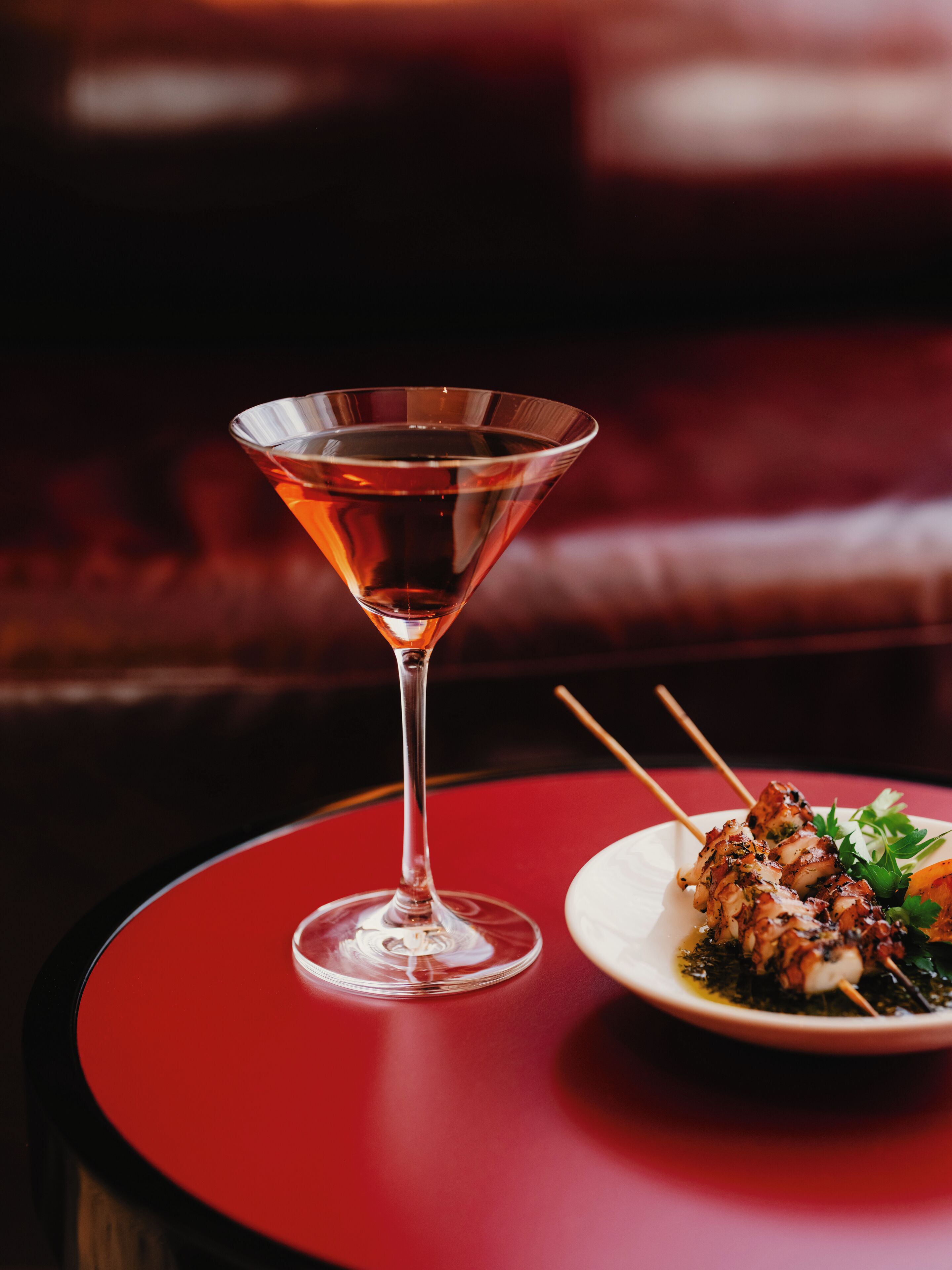 A martini glass on a red table with a plate of skewered food.