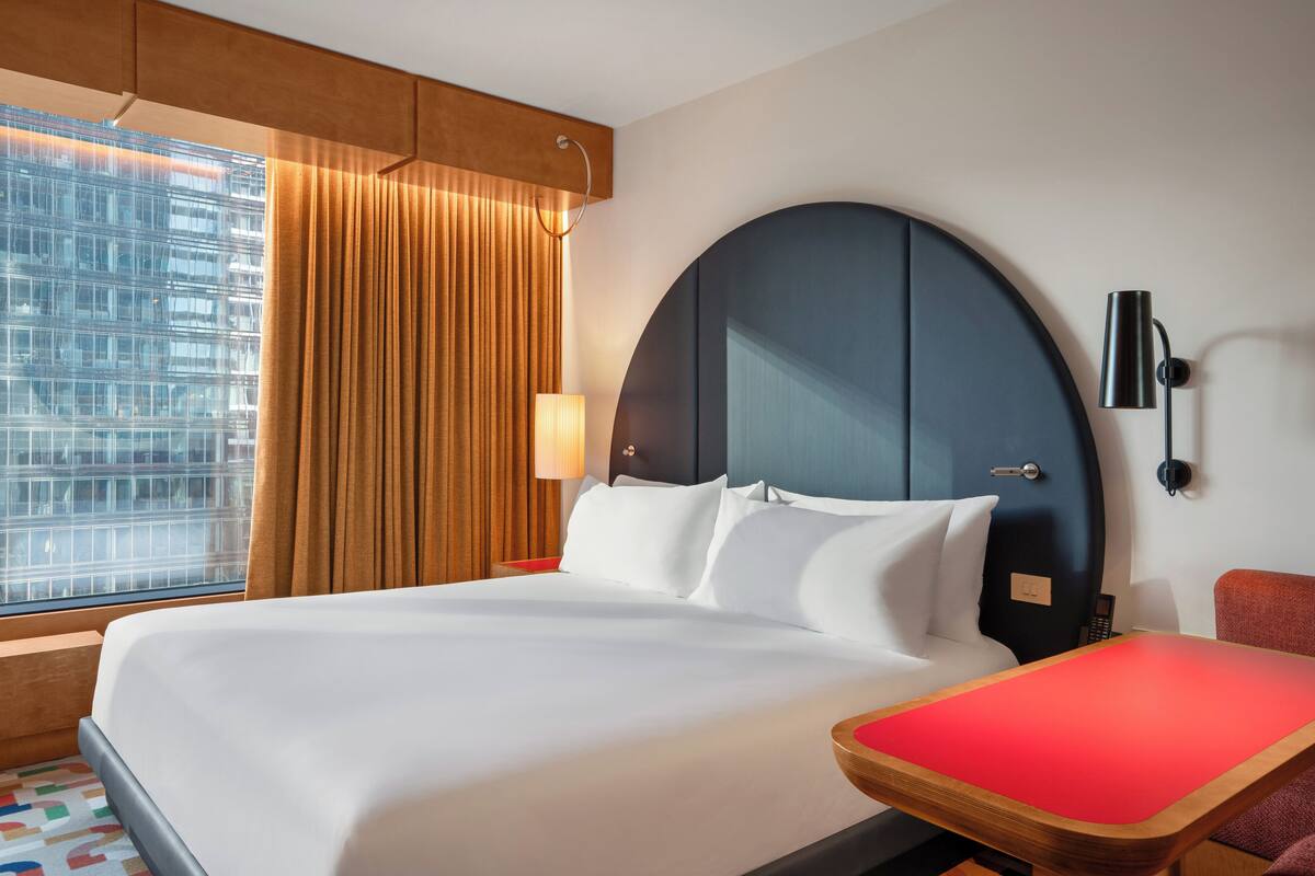 Habitación de hotel con una cama grande, un escritorio, una silla, una lámpara y vistas a un paisaje urbano por la ventana.