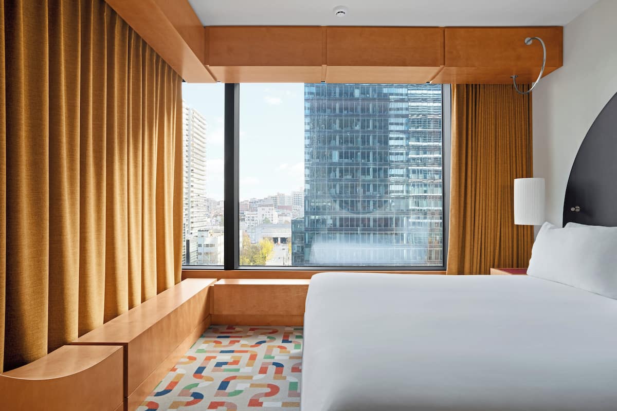 Habitación de hotel con un ventanal grande, una cama, una mesita de noche y vistas a un paisaje urbano.