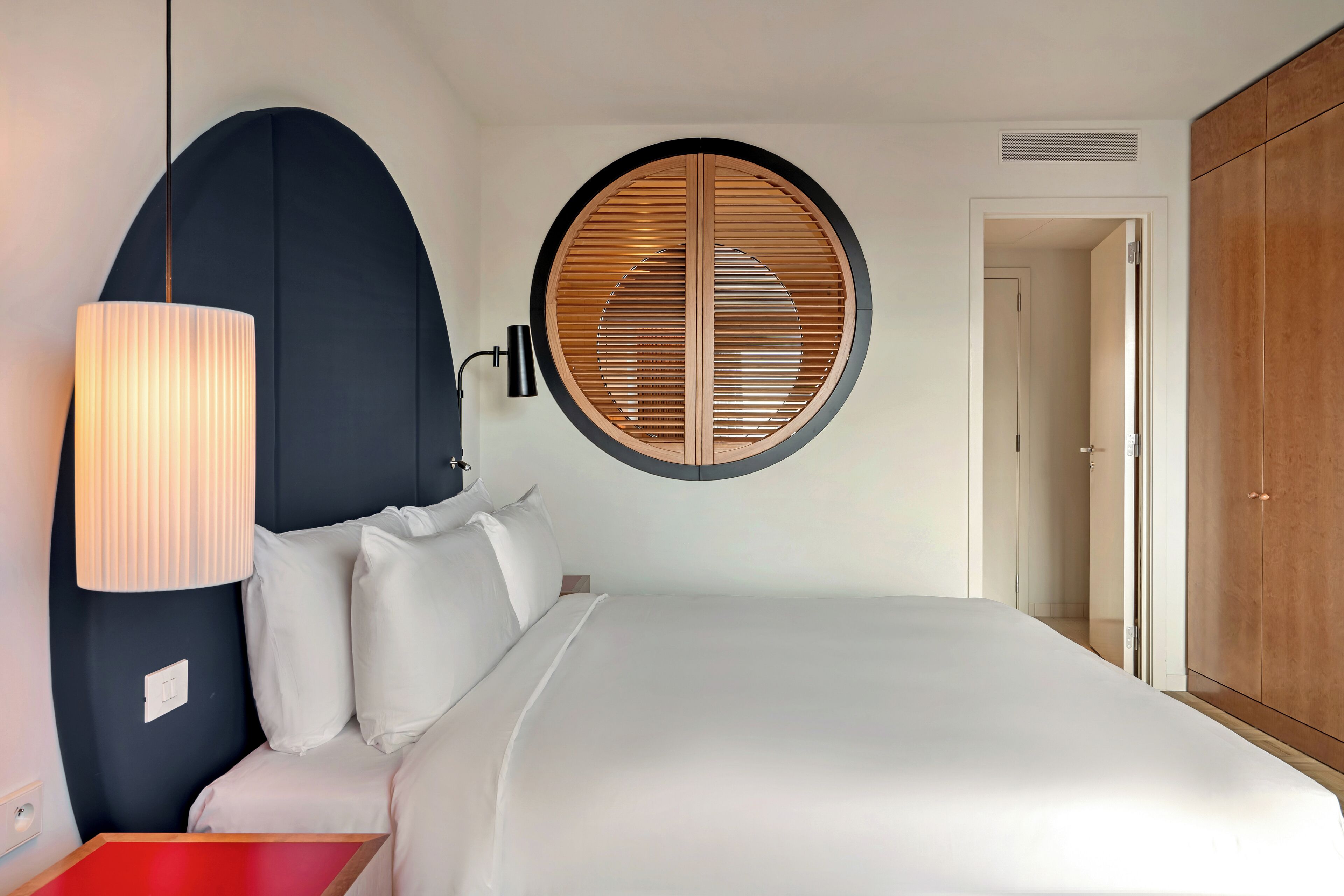 A neatly made bed with white linens, a round window with wooden blinds, a black lamp, and a wardrobe.