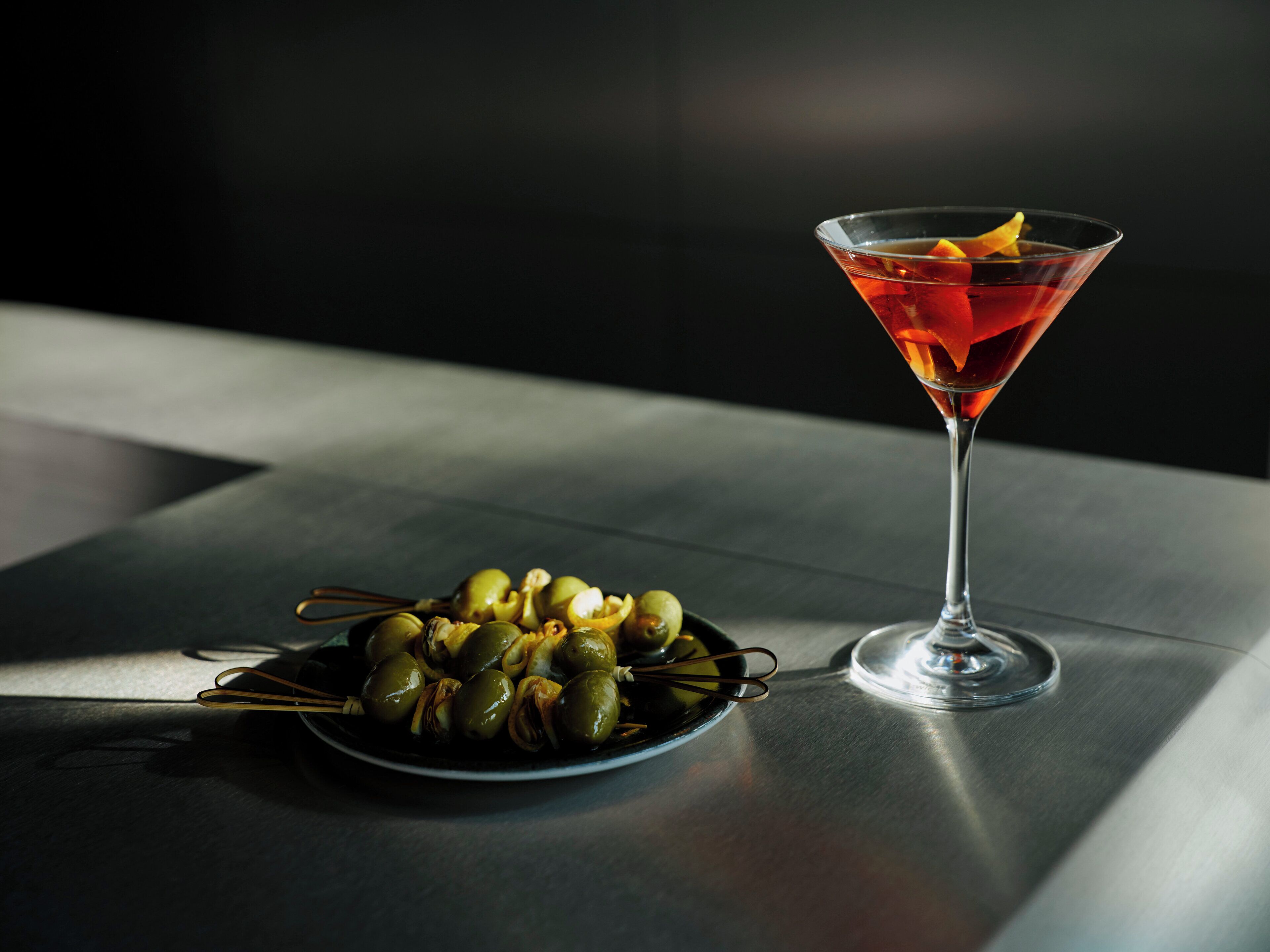 A martini glass with a lemon slice on a reflective surface, next to a plate of olives.