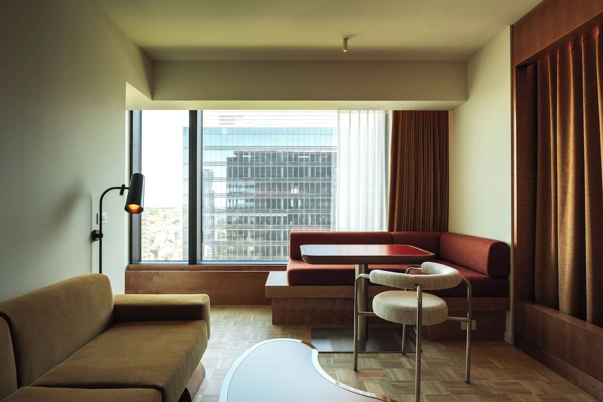 Una habitación de hotel moderna con sofá, una pequeña mesa redonda, una silla, un escritorio y un ventanal con cortinas.