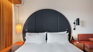 Una habitación de hotel moderna con una cama grande, un cabecero curvado, una mesita de noche con lámpara y un sofá rojo.