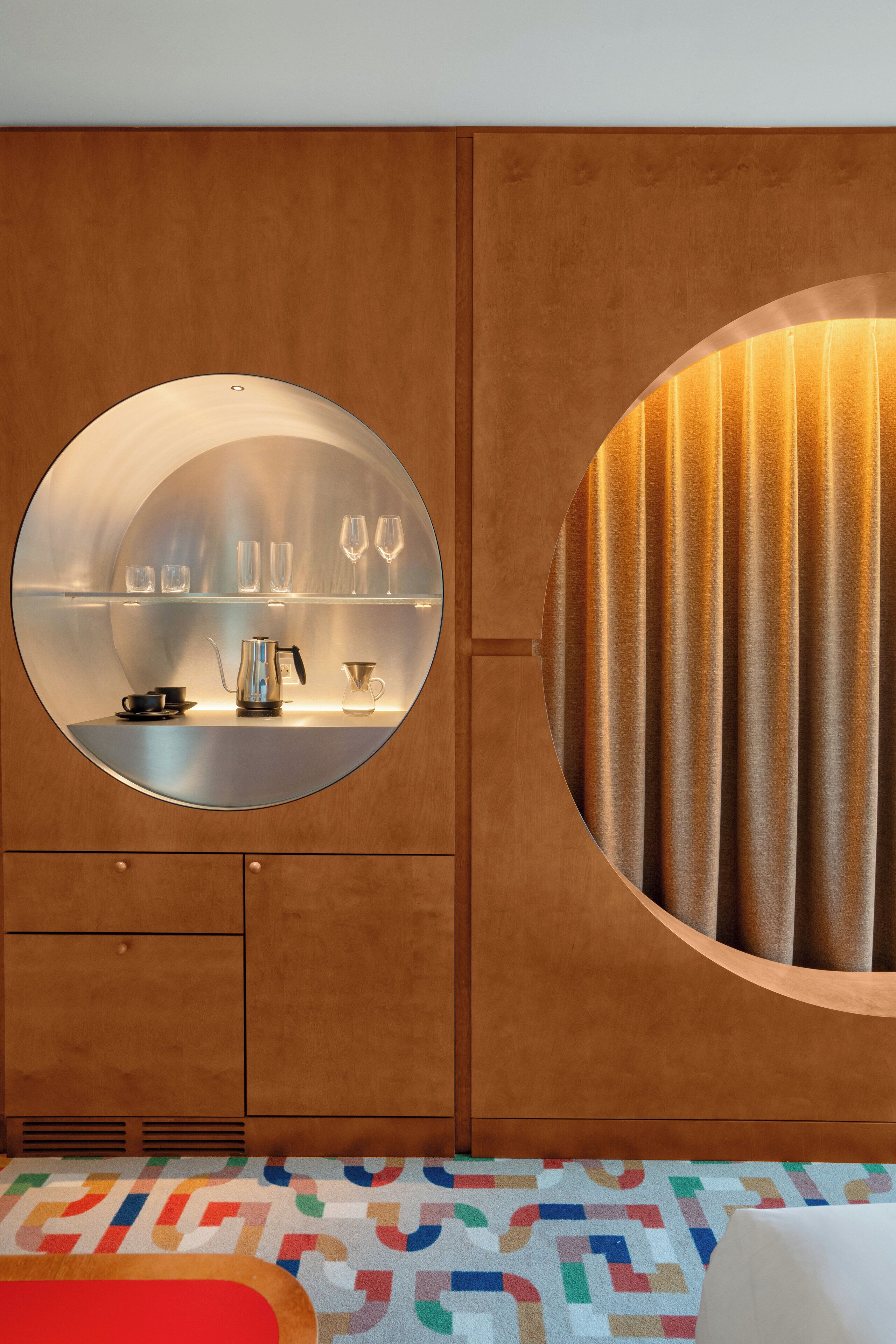 Una habitación con un espejo circular sobre un mueble de madera, una cama con un tapete de diseño geométrico y colores vivos, y un ventanal con cortinas.