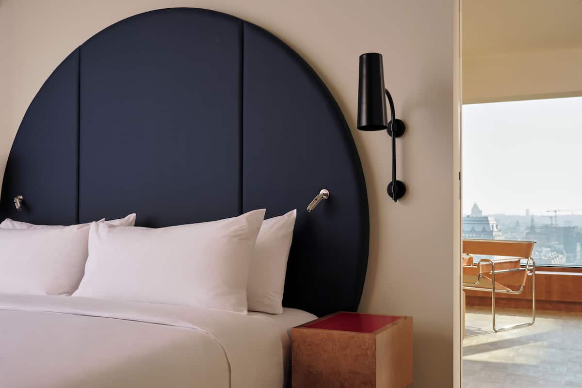 Una habitación de hotel moderna con una cama grande, cabecera redonda, mesita de noche y vistas a la ciudad a través de un ventanal amplio.