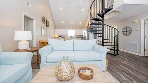 A spacious living room with a light blue sofa, a coffee table with decorative items, a staircase, and a kitchen in the background. - Bayside 3 bedroom Bungalow (Gulf Shores)