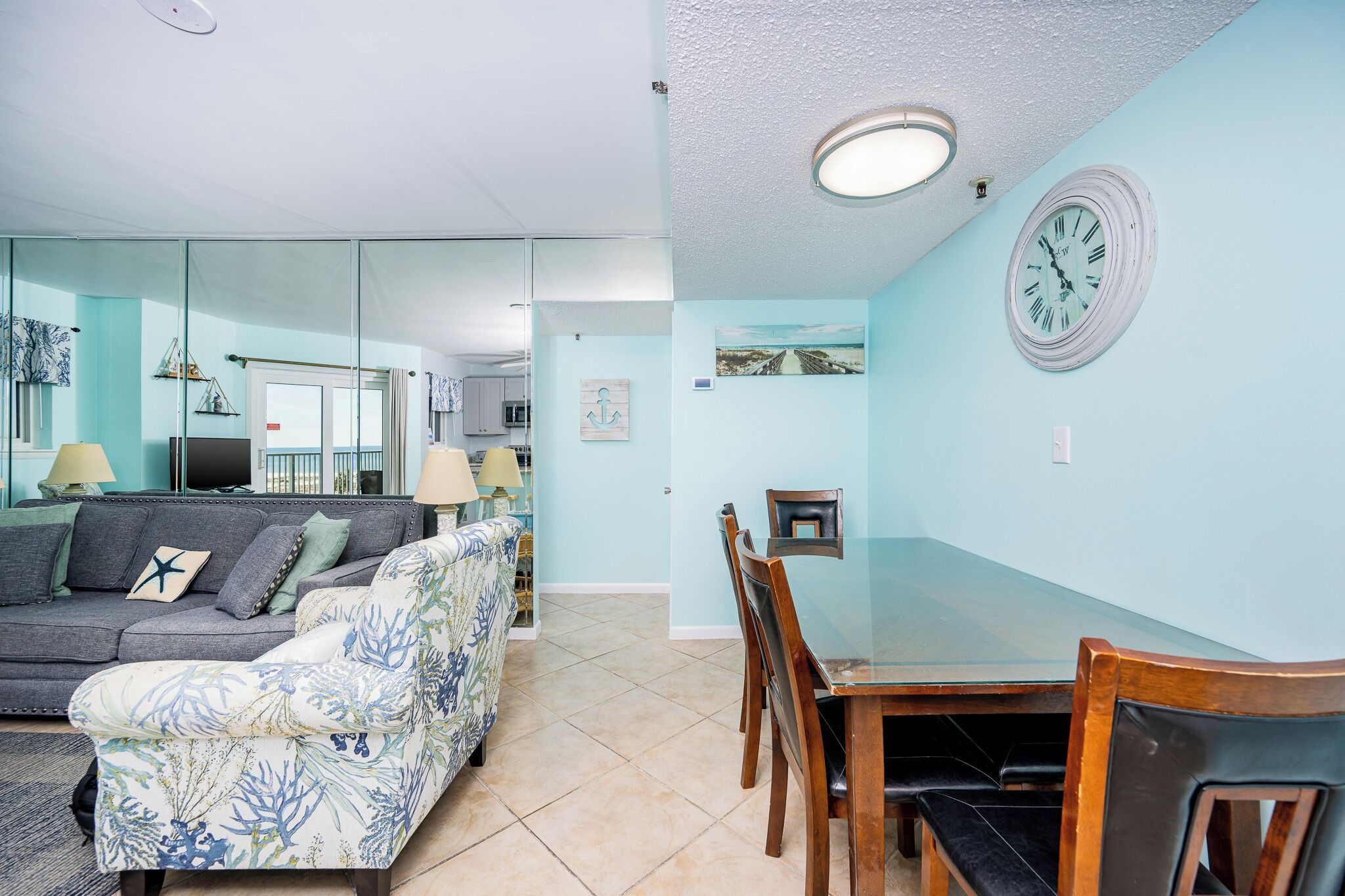 A living room with a sofa, a floral armchair, a dining table with chairs, and a wall-mounted clock.