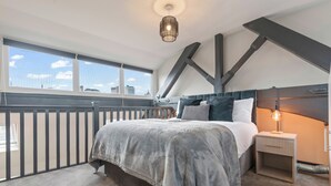 A bedroom with a large bed, a nightstand, a hanging light fixture, and a view of the city through a window.