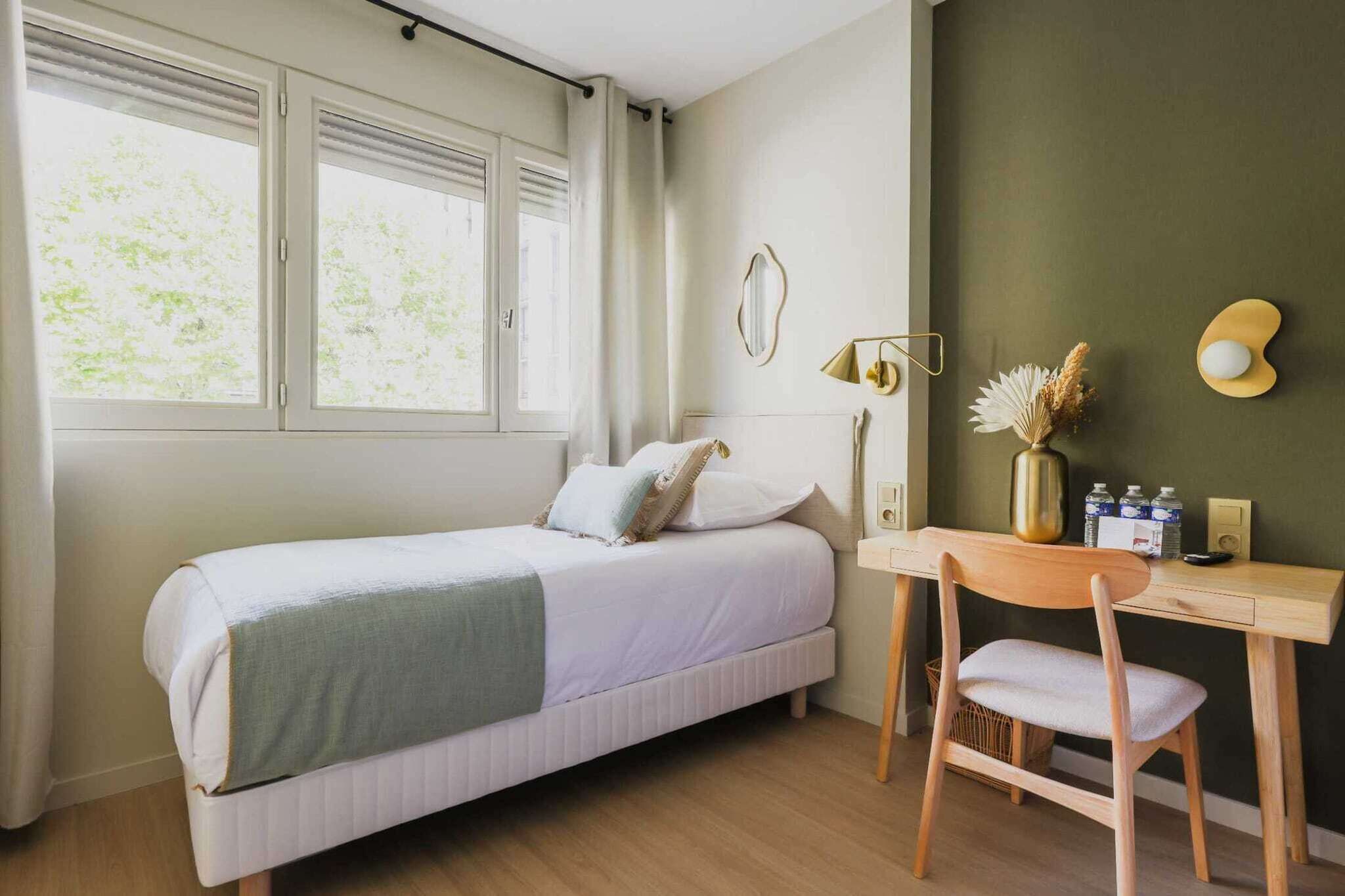 Une chambre à coucher avec un lit, un bureau et une chaise. Il y a une fenêtre avec des rideaux, une lampe fixée au mur et un vase contenant des fleurs séchées.