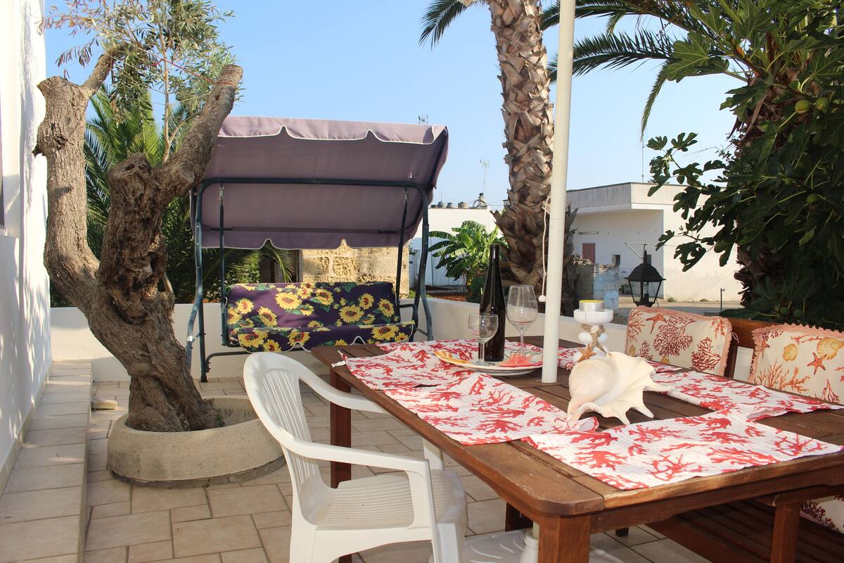 Une terrasse avec une table en bois, des chaises blanches, un espace salon abrité et un arbre.
