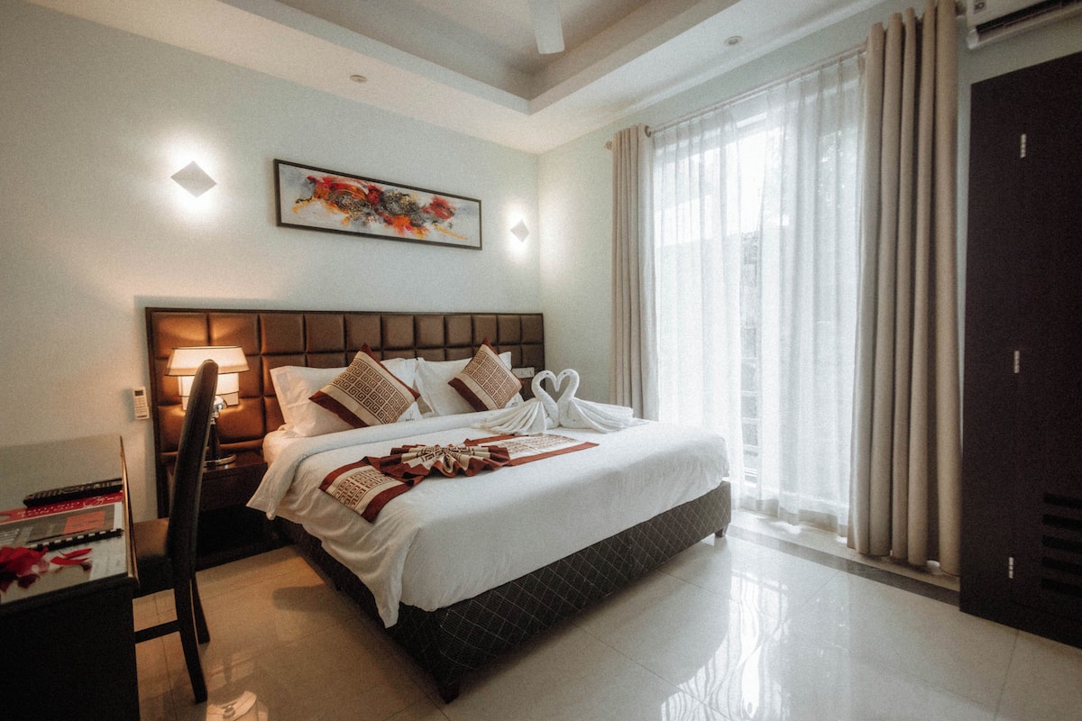 A hotel room with a large bed, a desk with a lamp, a chair, and a wardrobe.
