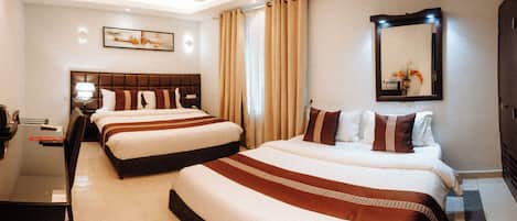 Une chambre d’hôtel avec deux lits, un bureau, une chaise, une lampe, un ventilateur de plafond et un grand miroir.