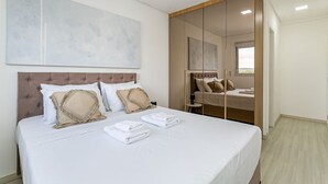 A neatly made bed with white linens and beige pillows, a large abstract painting above it, and a wooden wardrobe with a mirror.