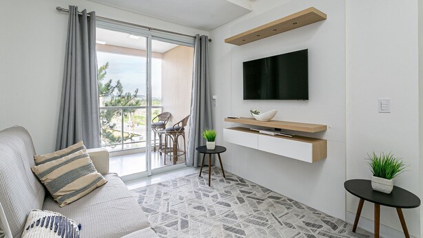 A modern living room with a sofa, a TV mounted on the wall, a small table with a plant, and a balcony with a view.