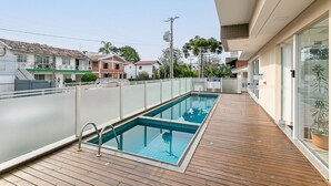 A swimming pool with a wooden deck, surrounded by a fence, and a clear view of the neighborhood.