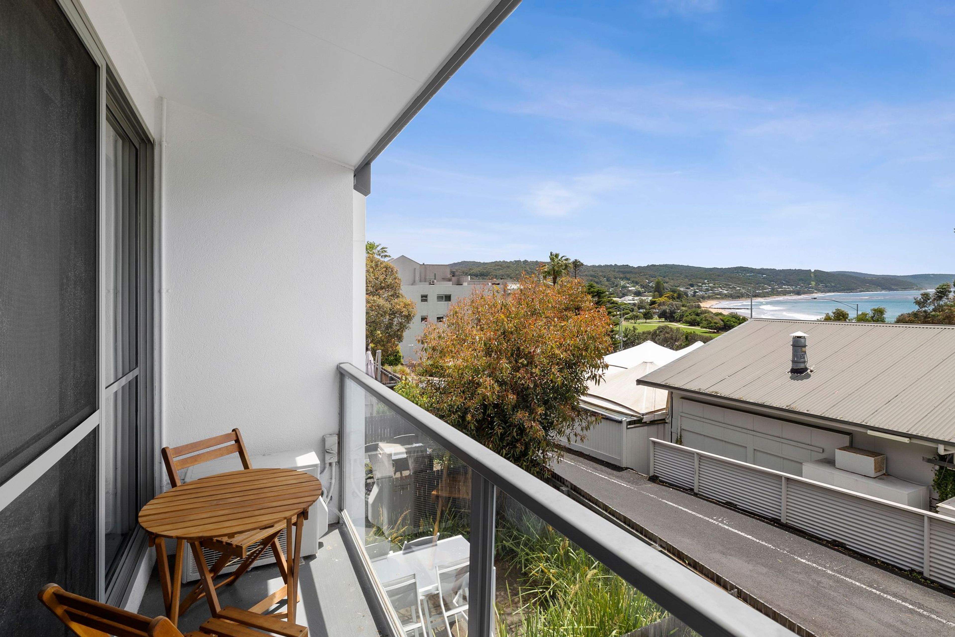 En balkon med et træbord og stole, der ud til et kystlandskab med bygninger og en klar himmel.