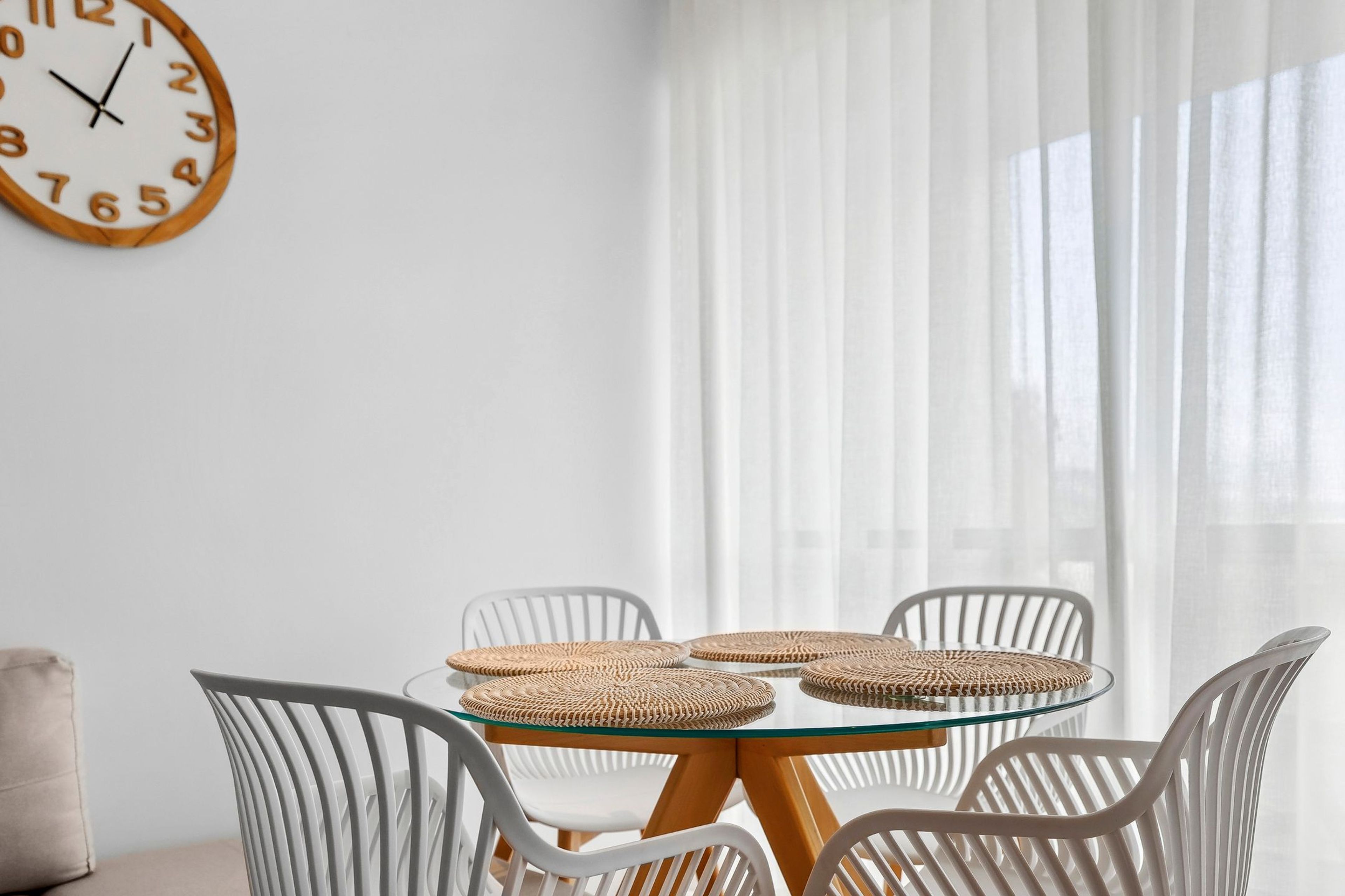 Een kamer met een ronde glazen tafel, omringd door witte stoelen, een geweven placemat en een houten klok aan de muur.