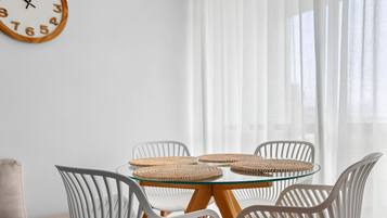 Een kamer met een ronde glazen tafel, omringd door witte stoelen, een geweven placemat en een houten klok aan de muur.