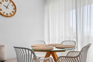 A room with a round glass table surrounded by white chairs, a woven placemat, and a wooden-framed clock on the wall.