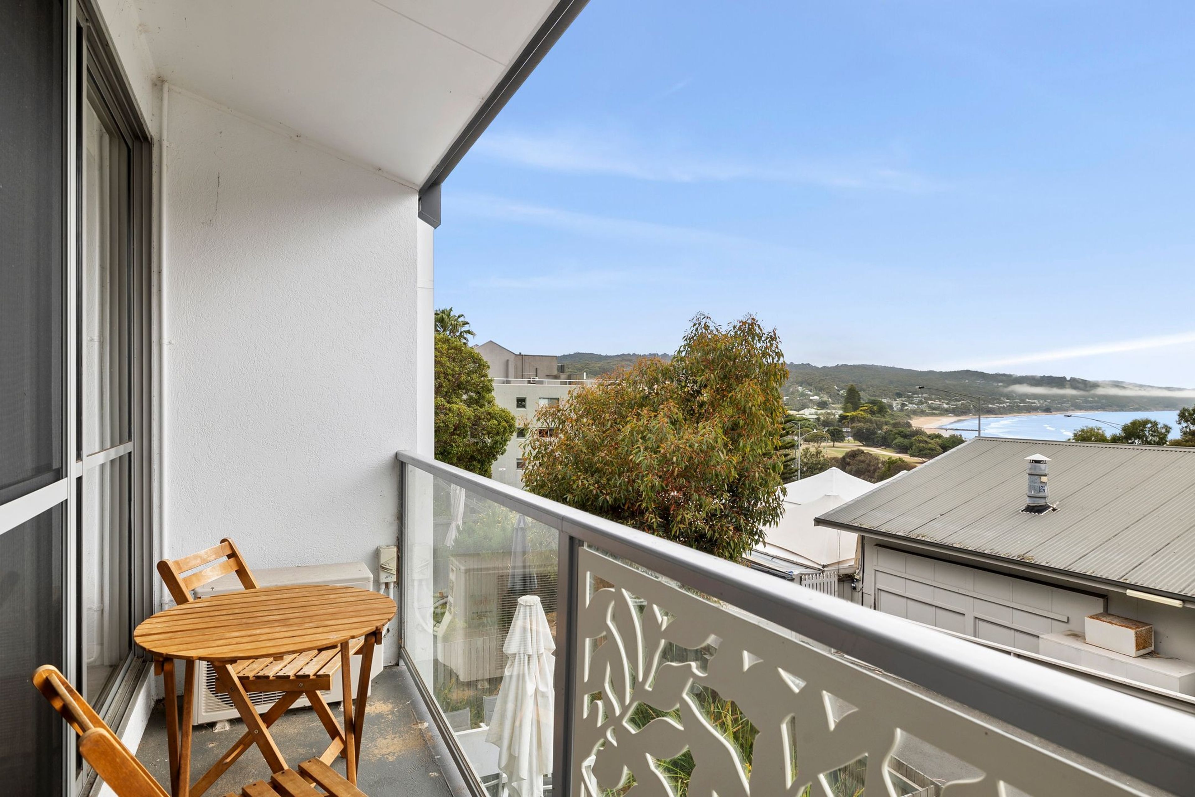 A balcony with a wooden table and chairs, overlooking a coastal landscape with buildings and trees.