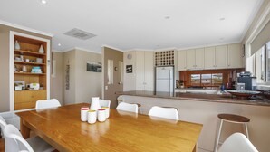 A modern kitchen with a central island, white cabinetry, and a dining area with a wooden table and white chairs. - Sound Of The Sea (Apollo Bay)