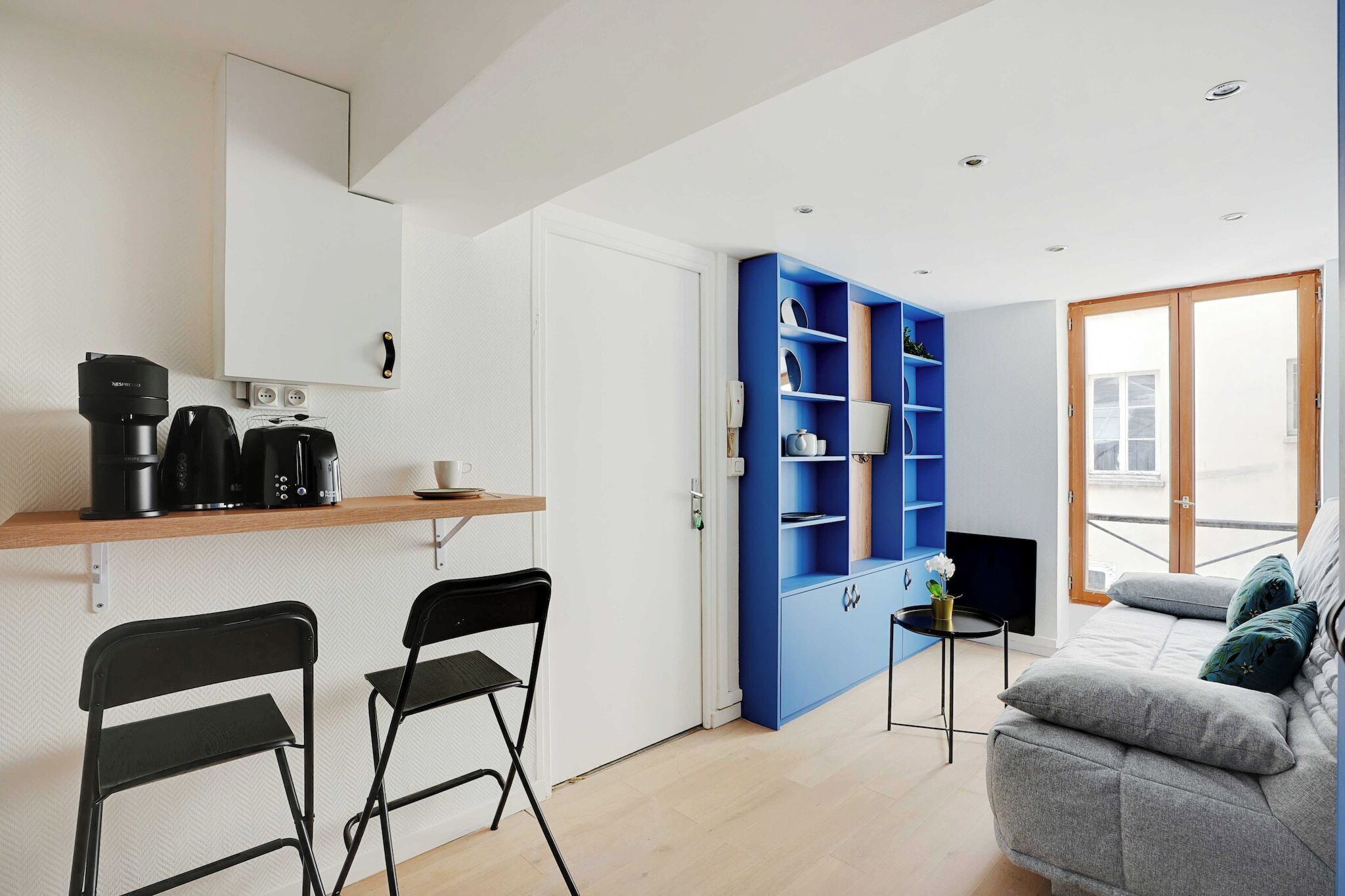 Een knus woongedeelte met een kitchenette, een blauwe wandplank, een grijze bank en een kleine tafel met een plant.