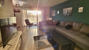 A modern living room with a sofa, a coffee table, a TV, and a chair. There is a view of the outdoors through a large window. - Luxury apartment (Lille)