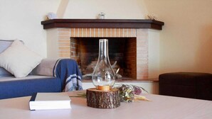 A living room with a brick fireplace, a clear glass vase on a wooden stump, a book, and a sofa with a cushion. - Villa Nerissa, Villa Nerissa (Alonnisos)