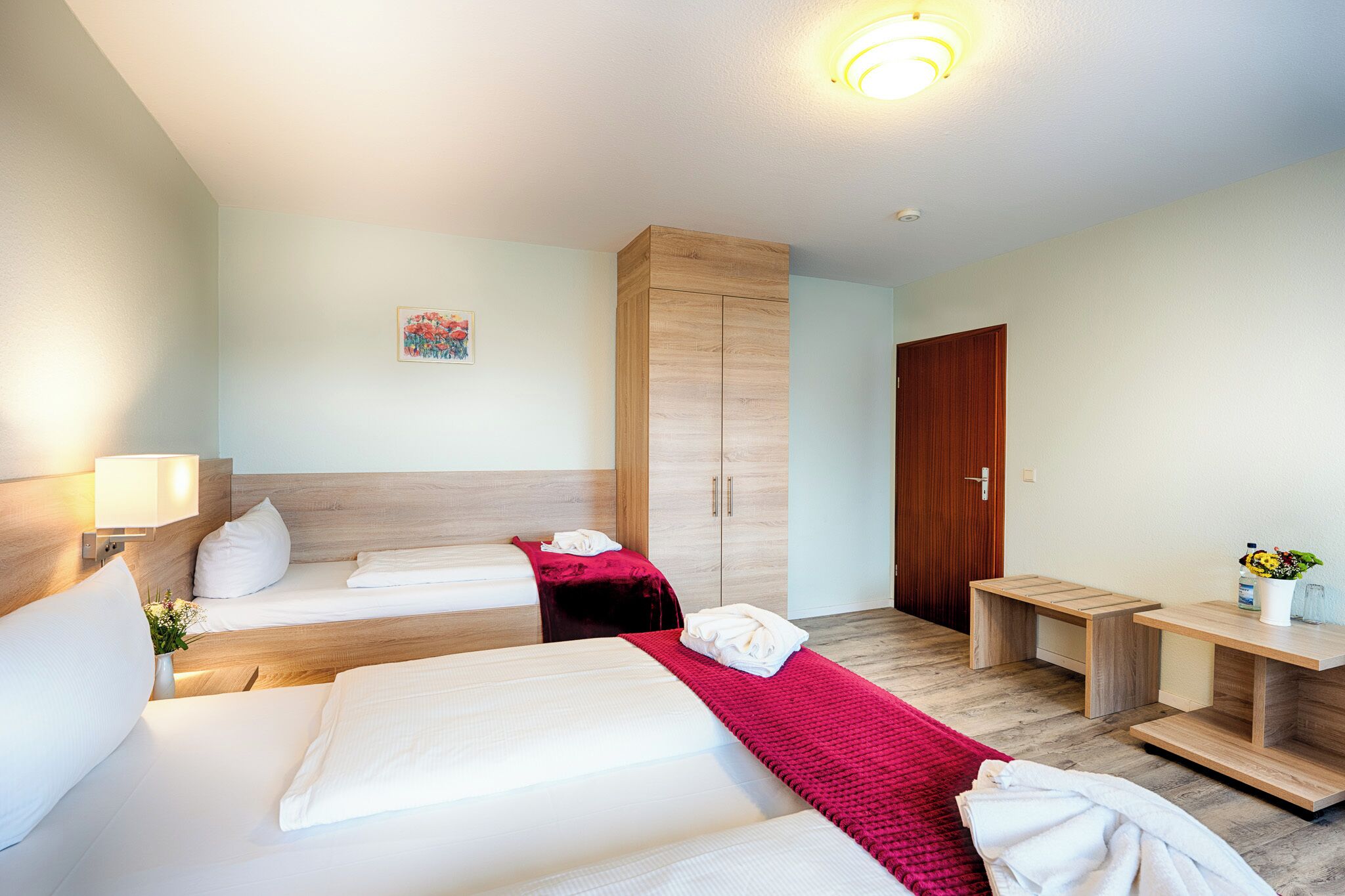 A hotel room with two beds, a wooden wardrobe, a desk, and a small table with a vase of flowers.