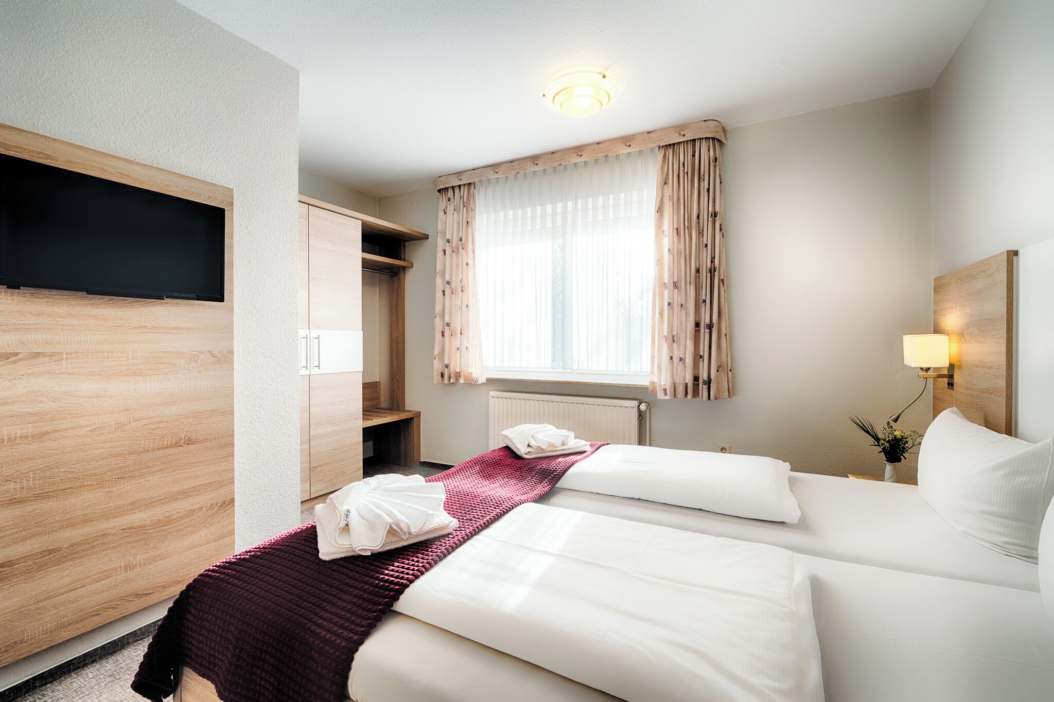 Ett hotellrum med två sängar, en tv som hänger på väggen, ett fönster med gardiner, en sänglampa och en liten växt på nattduksbordet.