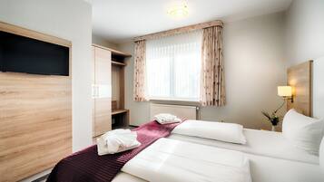 Ett hotellrum med två sängar, en tv som hänger på väggen, ett fönster med gardiner, en sänglampa och en liten växt på nattduksbordet.