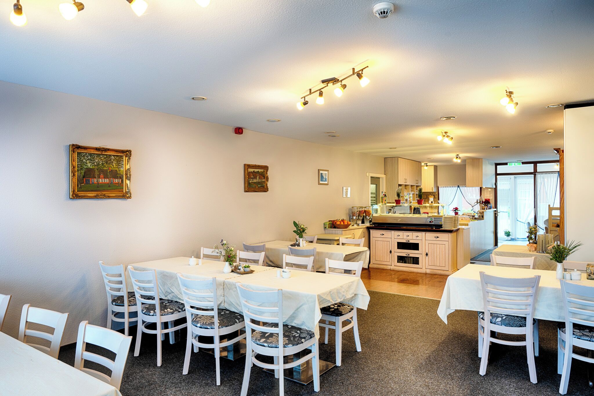 A dining area with multiple tables and chairs, a kitchen in the background, and ceiling lights.