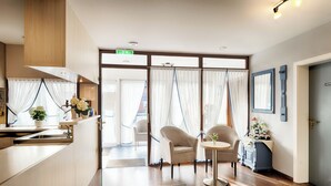 Quarto de hotel moderno com cozinha compacta, área de estar com duas cadeiras, uma mesa pequena e uma janela grande com cortinas leves.