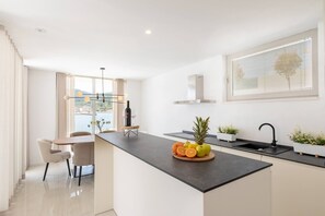 Une cuisine moderne dotée d’un îlot central, d’un coin repas avec des chaises, d’une fenêtre avec des stores et d’une vue sur l’extérieur.