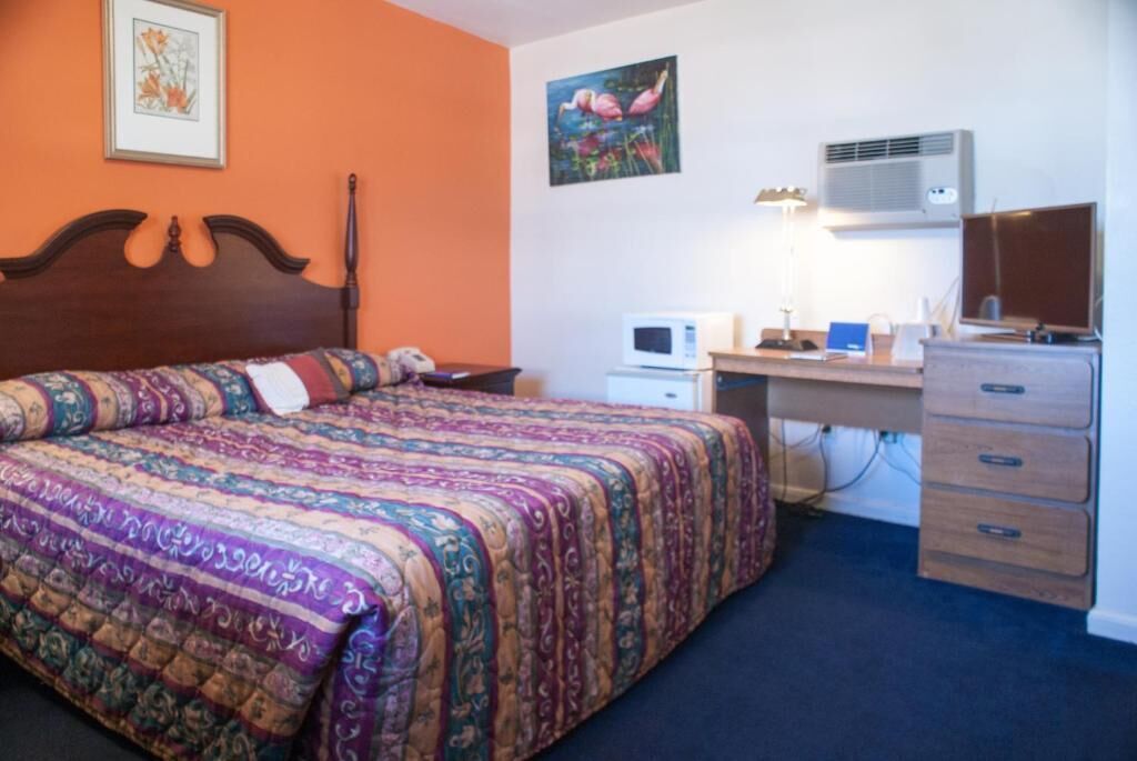 A bedroom with a large bed, a desk with a computer, a dresser, and an air conditioner.