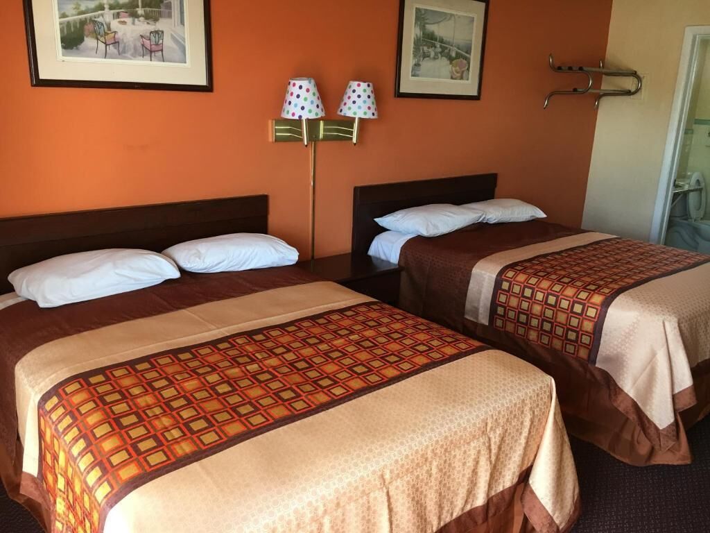 Hotellihuone, jossa on kaksi sänkyä. Jokaisessa sängyssä on kuviollinen päiväpeite ja valkoiset tyynyt. Seinälle on kiinnitetty kaksi lamppua, joissa on pistekuvioituja lampunvarjostimia.