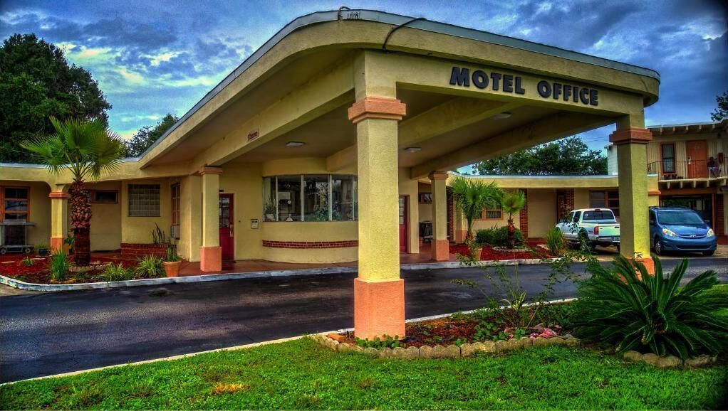 A motel office with a covered entrance, a parking area, and a small garden with palm trees.
