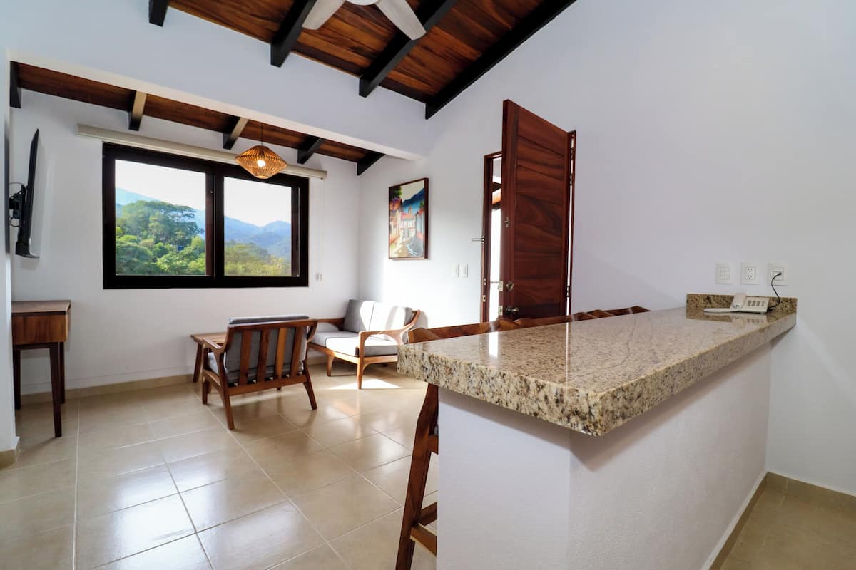 A kitchen with a granite countertop, wooden chairs, a dining table, a window with a view of greenery, and a ceiling with exposed beams.