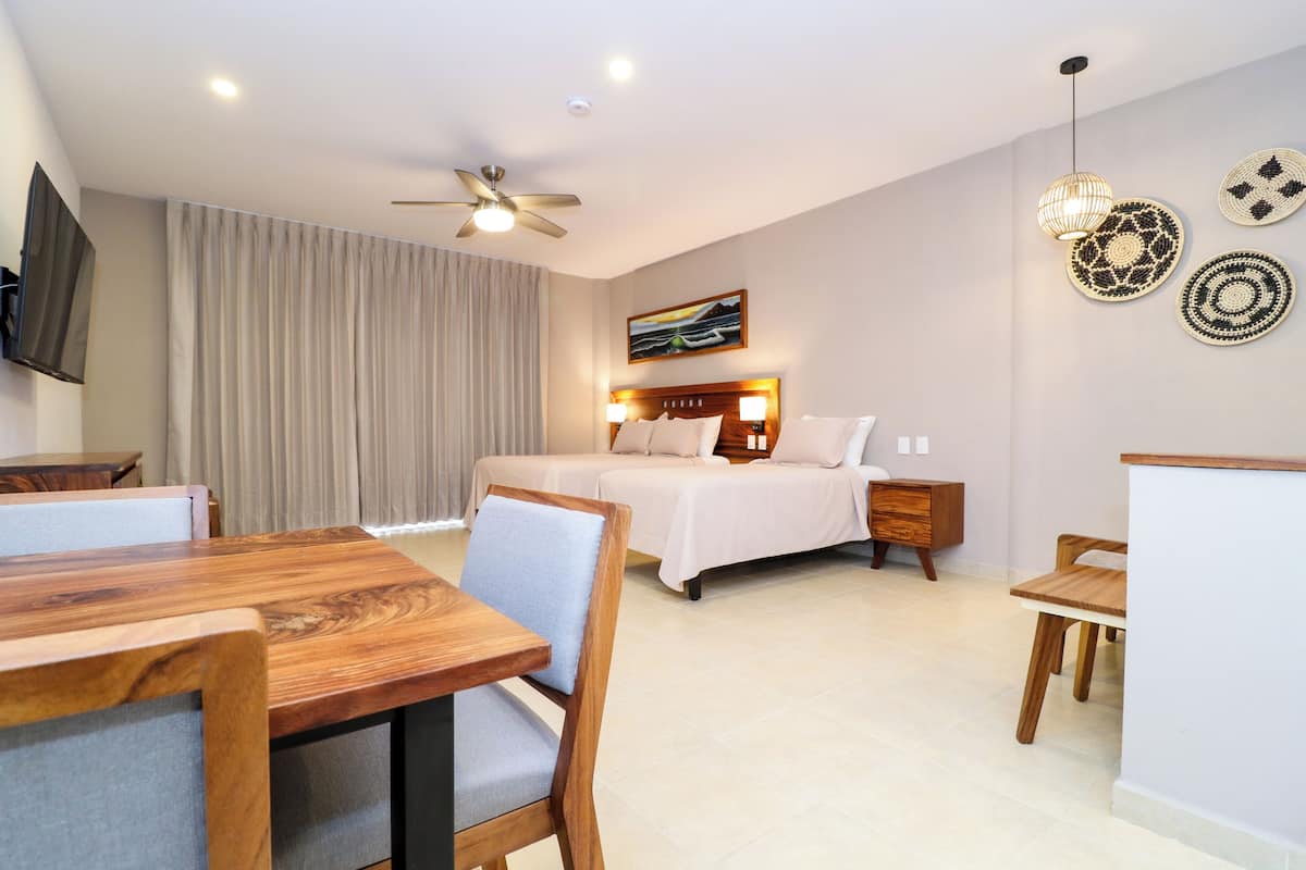 A hotel room with two beds, a dining area with wooden furniture, a ceiling fan, and wall-mounted artwork.