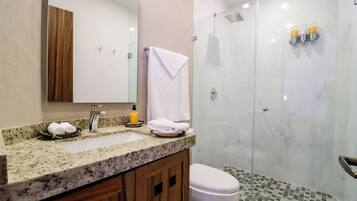 Un baño con encimera de granito, un lavabo blanco, un espejo, un mueble de madera, un inodoro blanco y una zona de ducha con mampara de cristal.