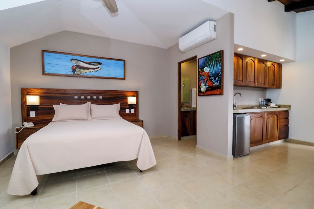 A bedroom with a bed, a nightstand, a painting, and a view into the kitchen.