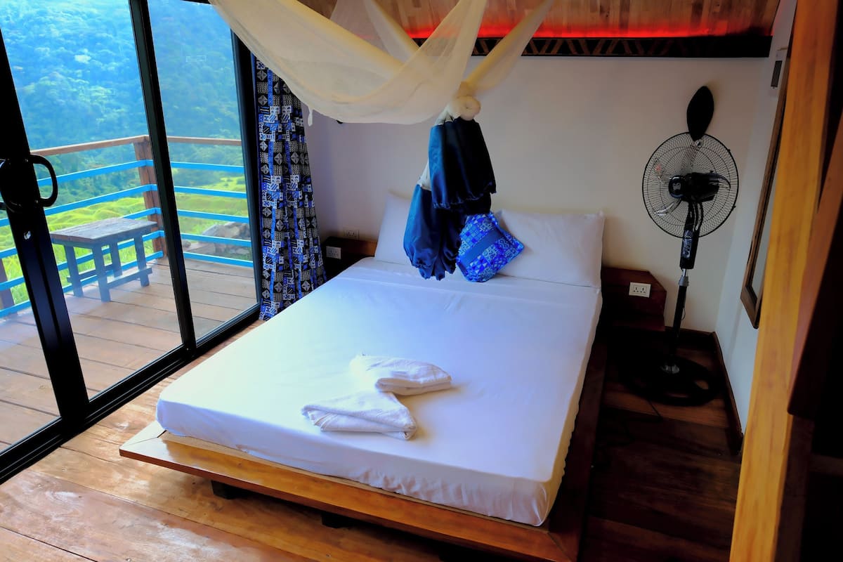 Quarto com cama, ventilador de cabeceira e vista para um deck e guarda-corpo de madeira.