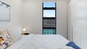 Een slaapkamer met een bed, nachtkastje, lamp, raam met jaloezieën en een kledingkast.