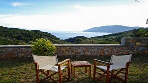 Une terrasse avec deux chaises en bois et une table, d’où l’on peut admirer un paysage côtier avec des collines et la mer.